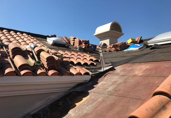 The image shows a roof with damaged tiles and debris scattered around, under a clear blue sky.