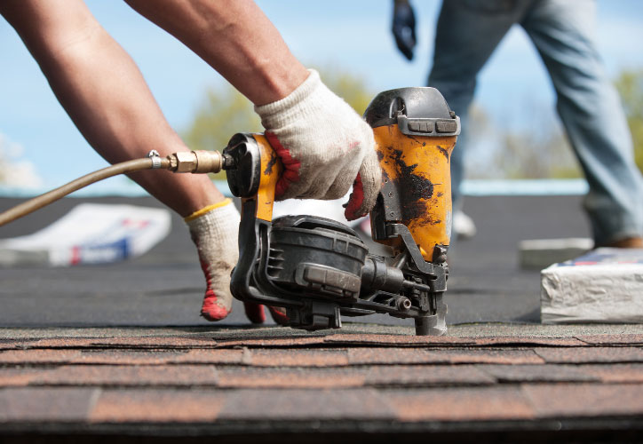 A construction worker using a power drill on a roofing project with other workers in the background.