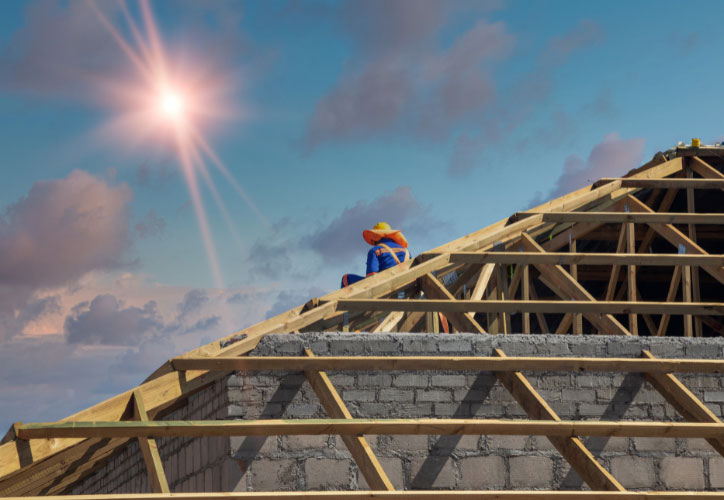 The image shows a construction site with a partially built house, featuring an unfinished roof, under a clear sky during daylight hours.