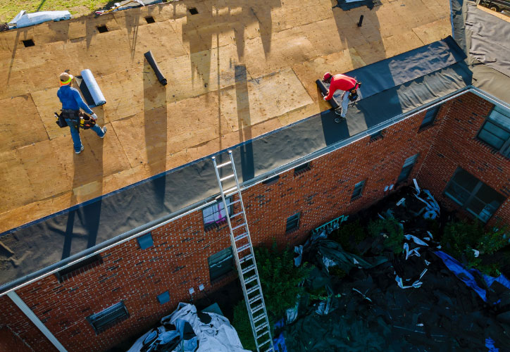 The image shows two individuals working on a rooftop construction project, with one person standing on the roof and another standing on a ladder, both wearing safety gear, under a clear sky.