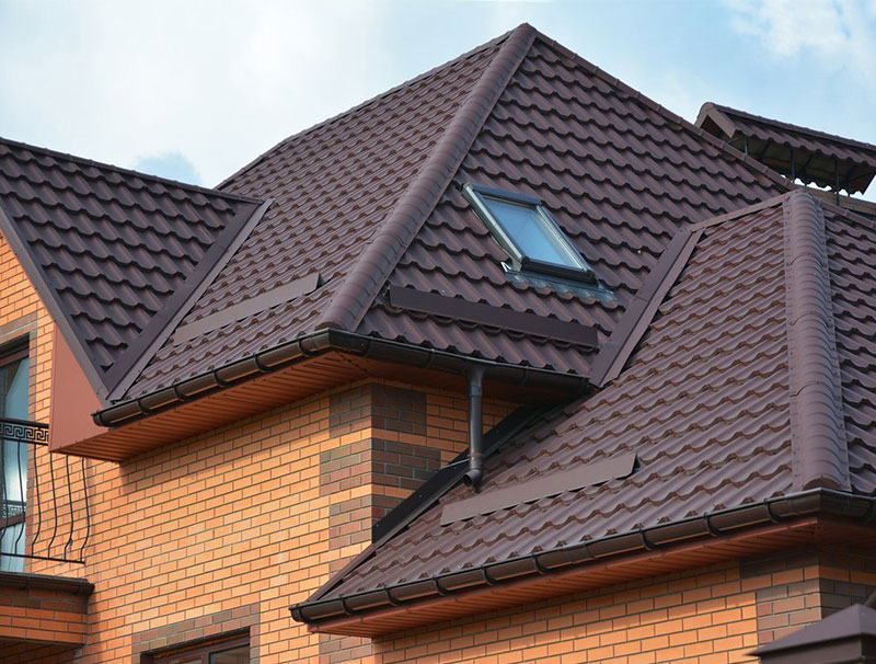 The image shows a residential house with a pitched roof featuring brown shingles and a prominent gable with a skylight, surrounded by trees and sky.